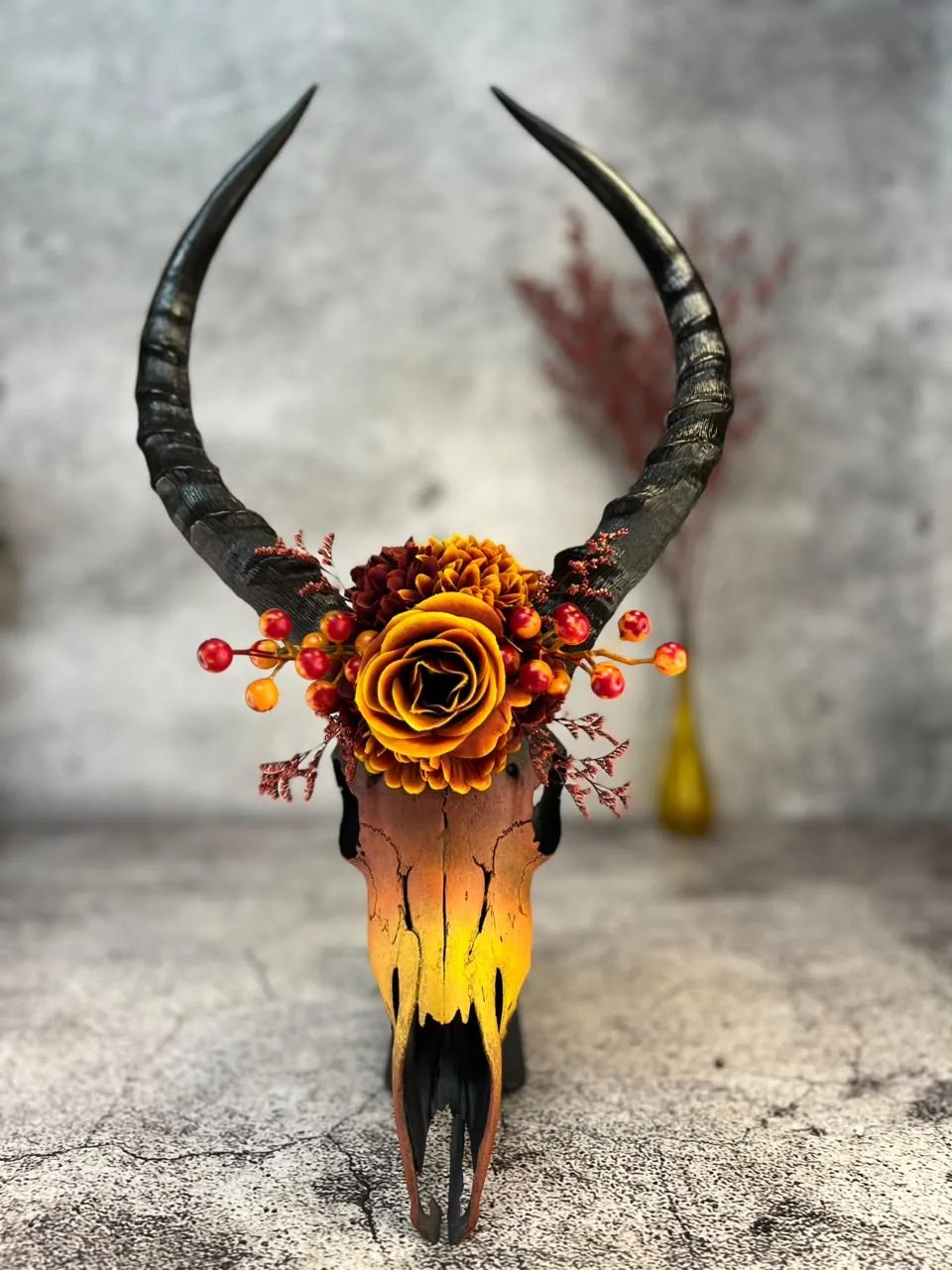 Skull painted in a warm orange-yellow gradient, crowned with autumn roses and berries between curved black horns